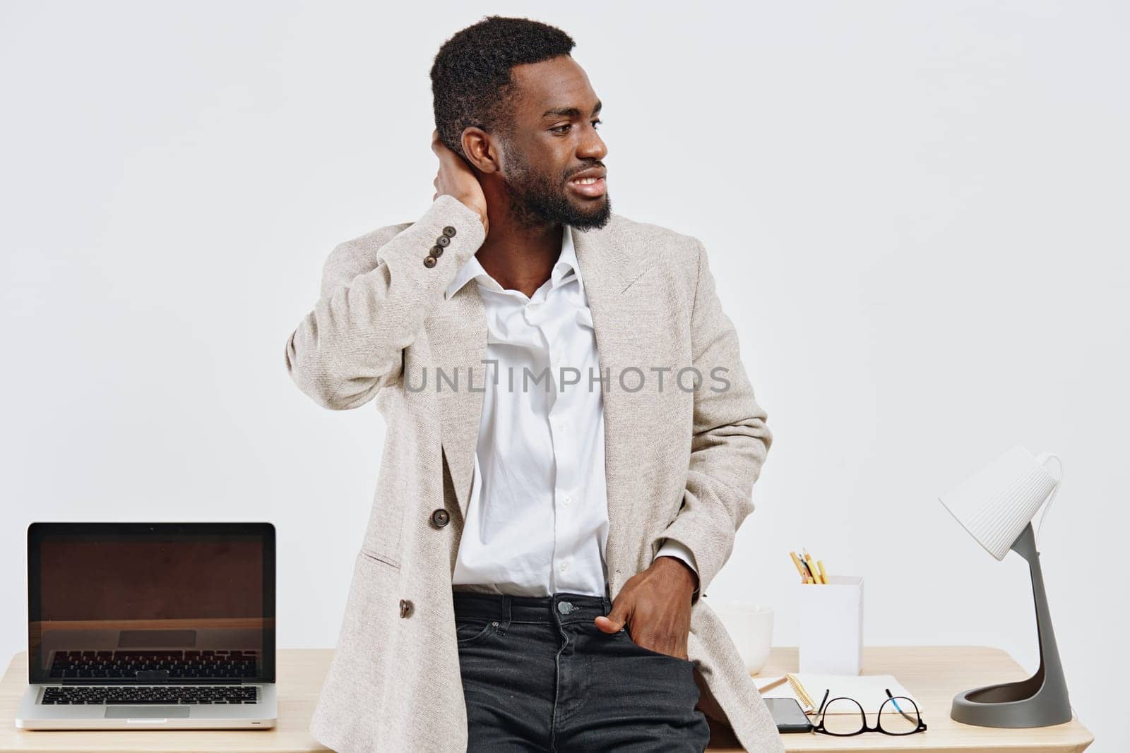Young professional man with dark curly hair, wearing a beige blazer and white shirt, standing in a contemporary office environment and smiling. He appears relaxed and confident, with a laptop, glasses, and office supplies on the desk. Concept: workplace success.
