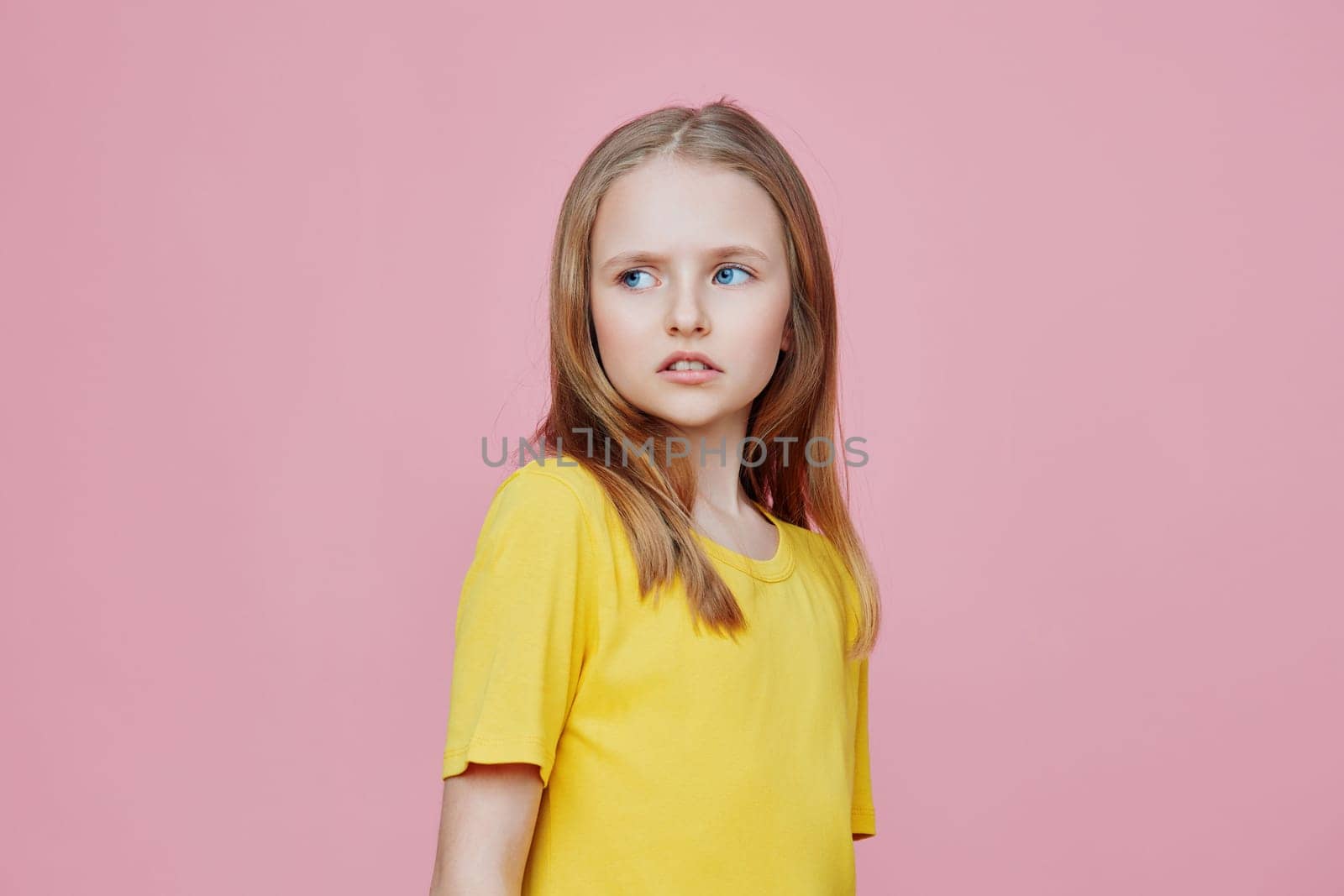 A young girl wearing a yellow shirt poses against a soft pink background, showcasing her thoughtful expression and innocence, perfect for themes of childhood and emotion