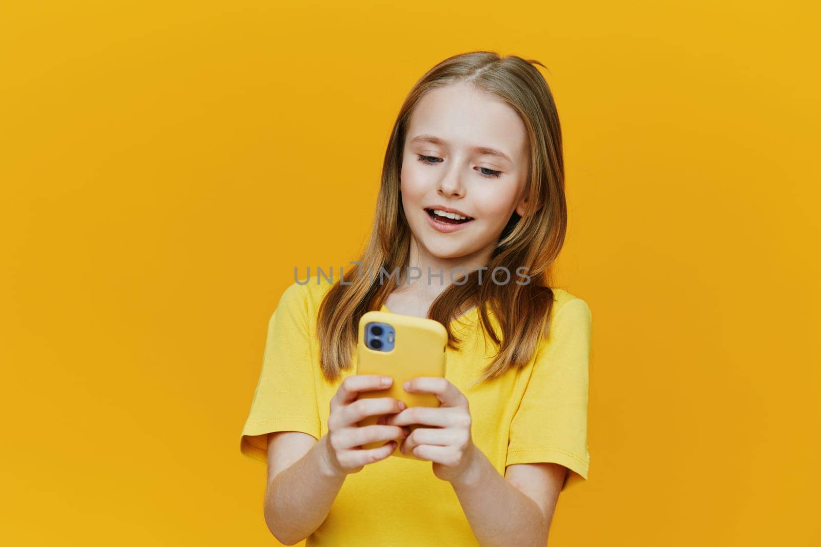 Cheerful girl in yellow shirt smiles while using her smartphone with a matching yellow case, set against a lively yellow backdrop, capturing the essence of youthful joy and modern technology