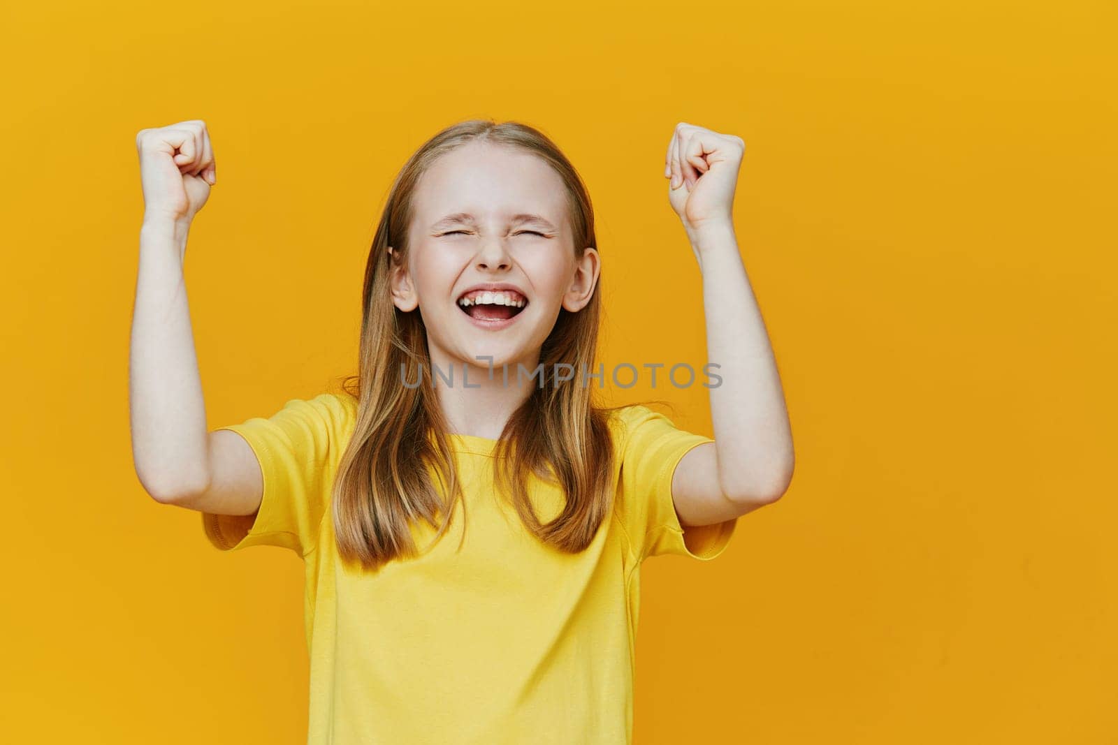 A joyful young girl expressing happiness and excitement, wearing a yellow t shirt, with raised arms against a vibrant yellow background, symbolizing success and positivity