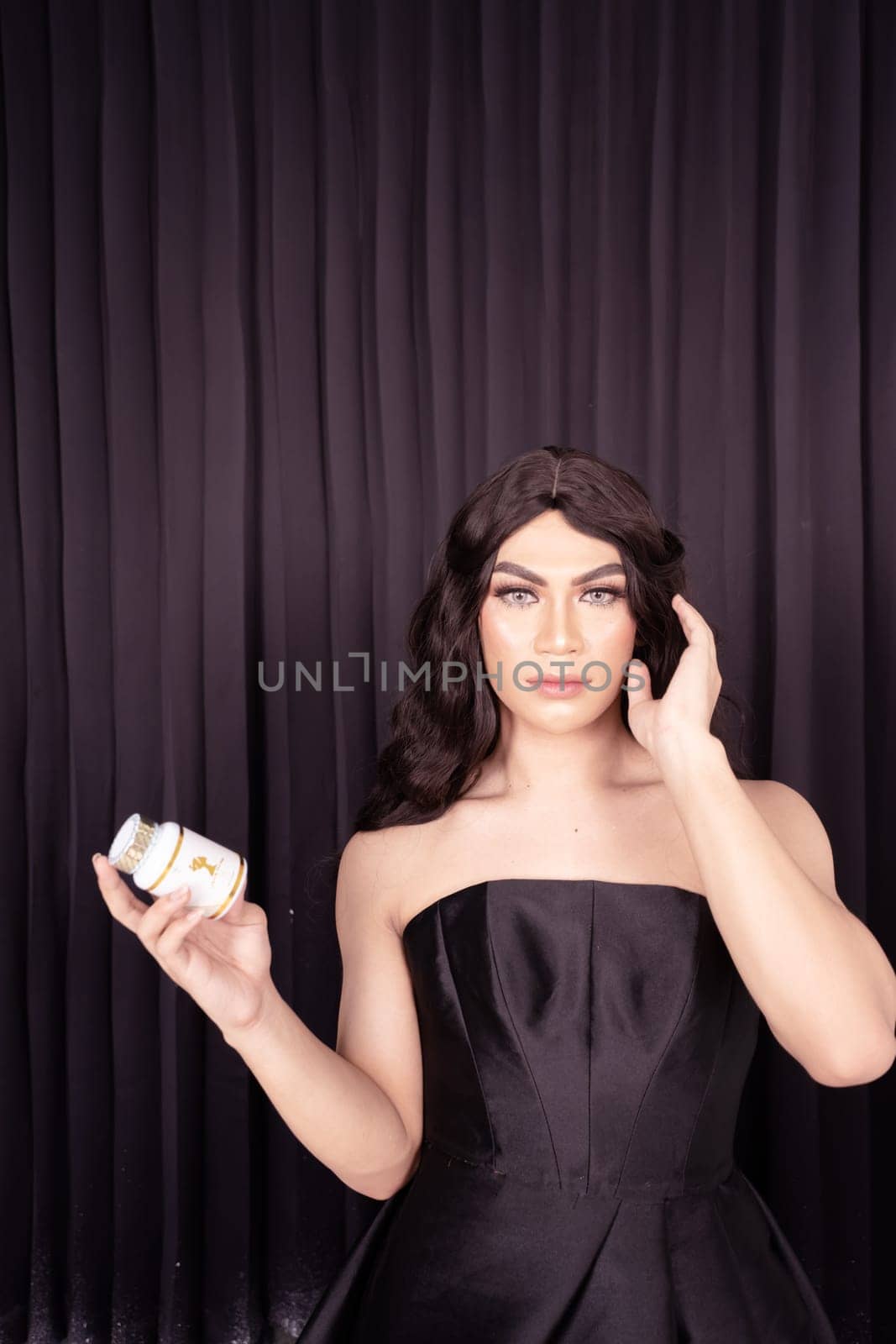 Beautiful Woman holding skincare in her hand while wearing a black dress with black hair in front of the black curtain