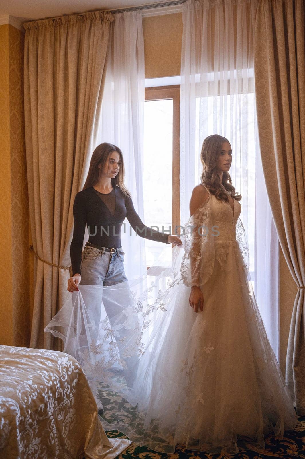 A bridesmaid helps a young bride get ready for her wedding day. A bridesmaid adjusts an elegant bride's lace dress. The morning of the wedding day.