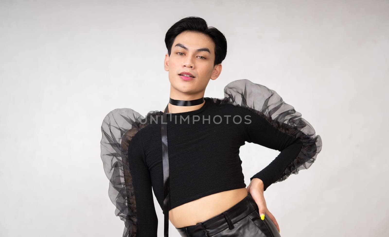 Portrait of a feminine man in black dress and black hair while wearing makeup inside the white studio