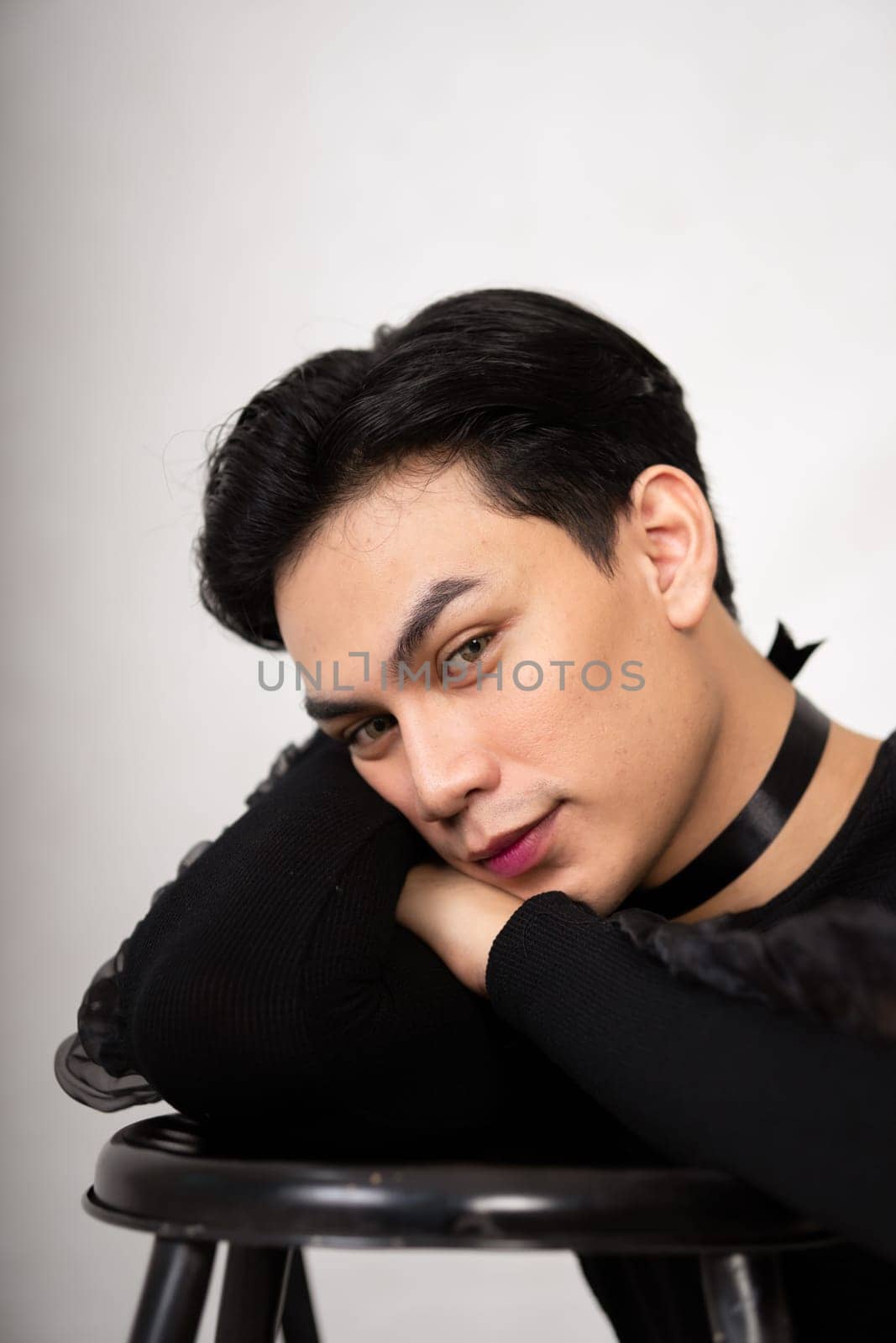 Pretty Asian man putting his hand and head on the chair while posing in a black dress inside the studio