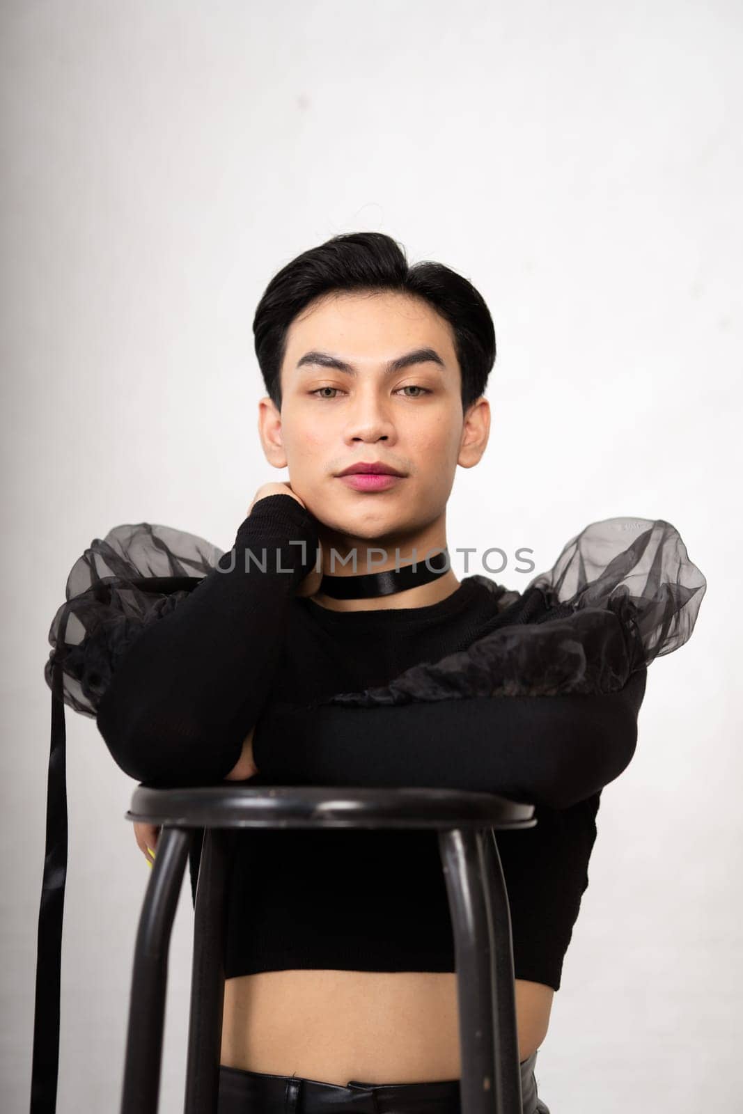 beautiful Asian man posing with the chair while wearing a black costume and makeup inside the white studio