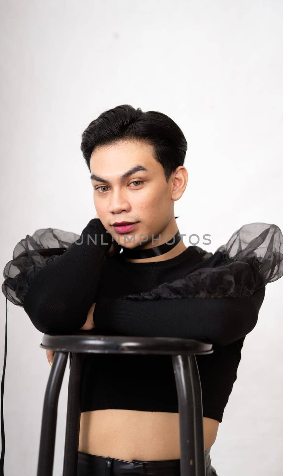 beautiful Asian man posing with the chair while wearing a black costume and makeup inside the white studio