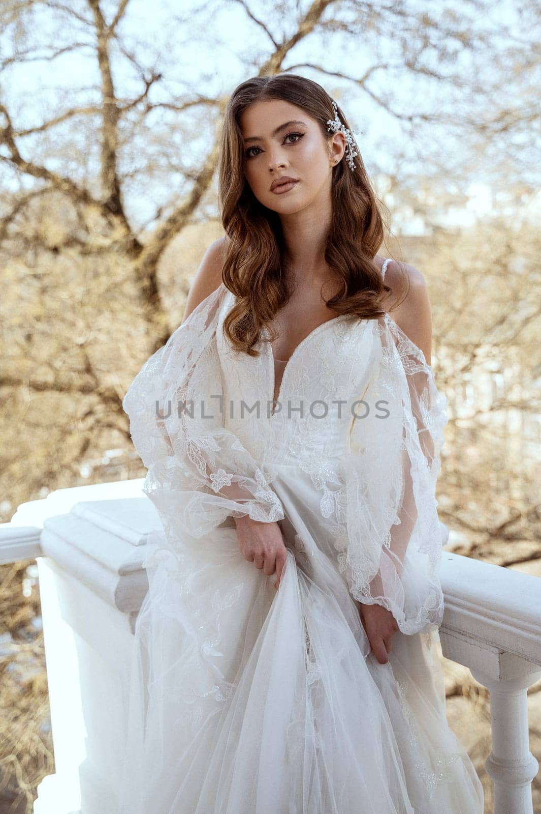 Young brunette bride in traditional long white dress posing on hotel balcony in spring. white dress with long sleeves, corset and open shoulders. Bride portrait, wedding makeup, fashionable bride.