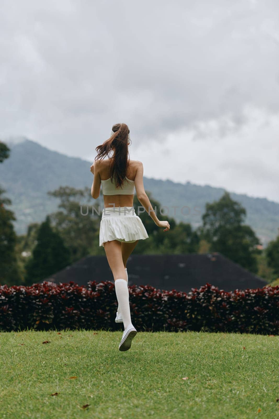 A young woman wearing a white athletic ensemble runs on lush grass, displaying a sense of freedom and energy in a scenic outdoor backdrop, emphasizing fitness and well being