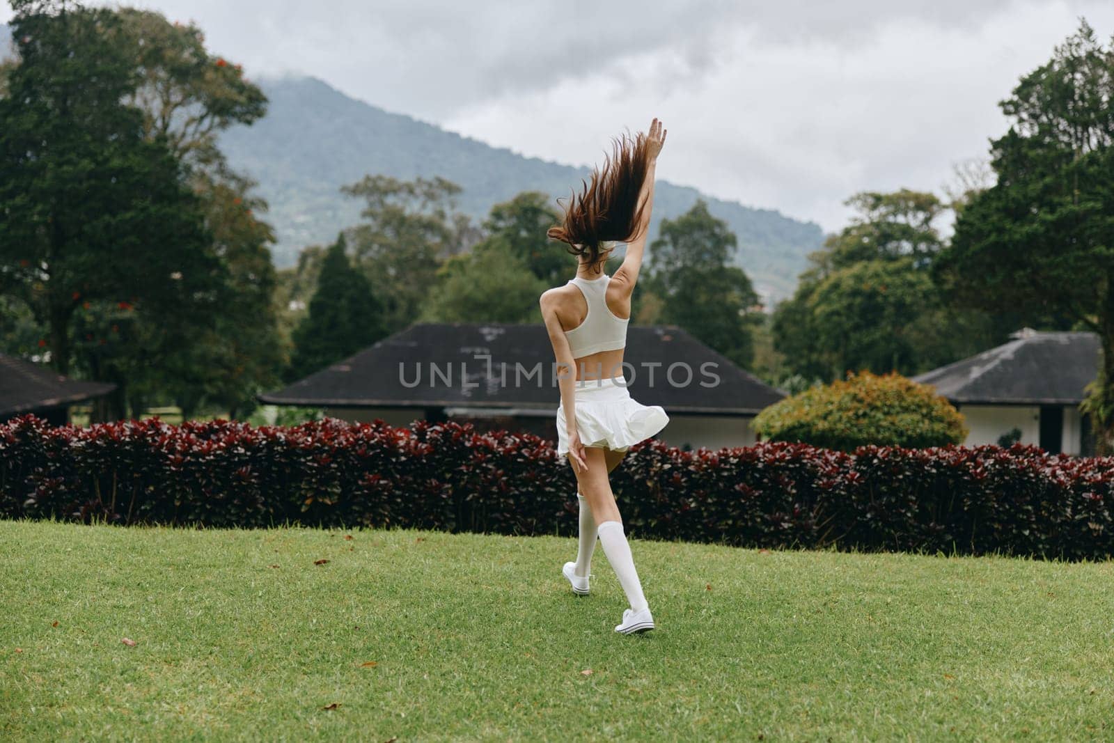 A young woman in a white sports outfit, dancing happily in a vibrant green park with mountains behind, embodying joy and freedom in nature