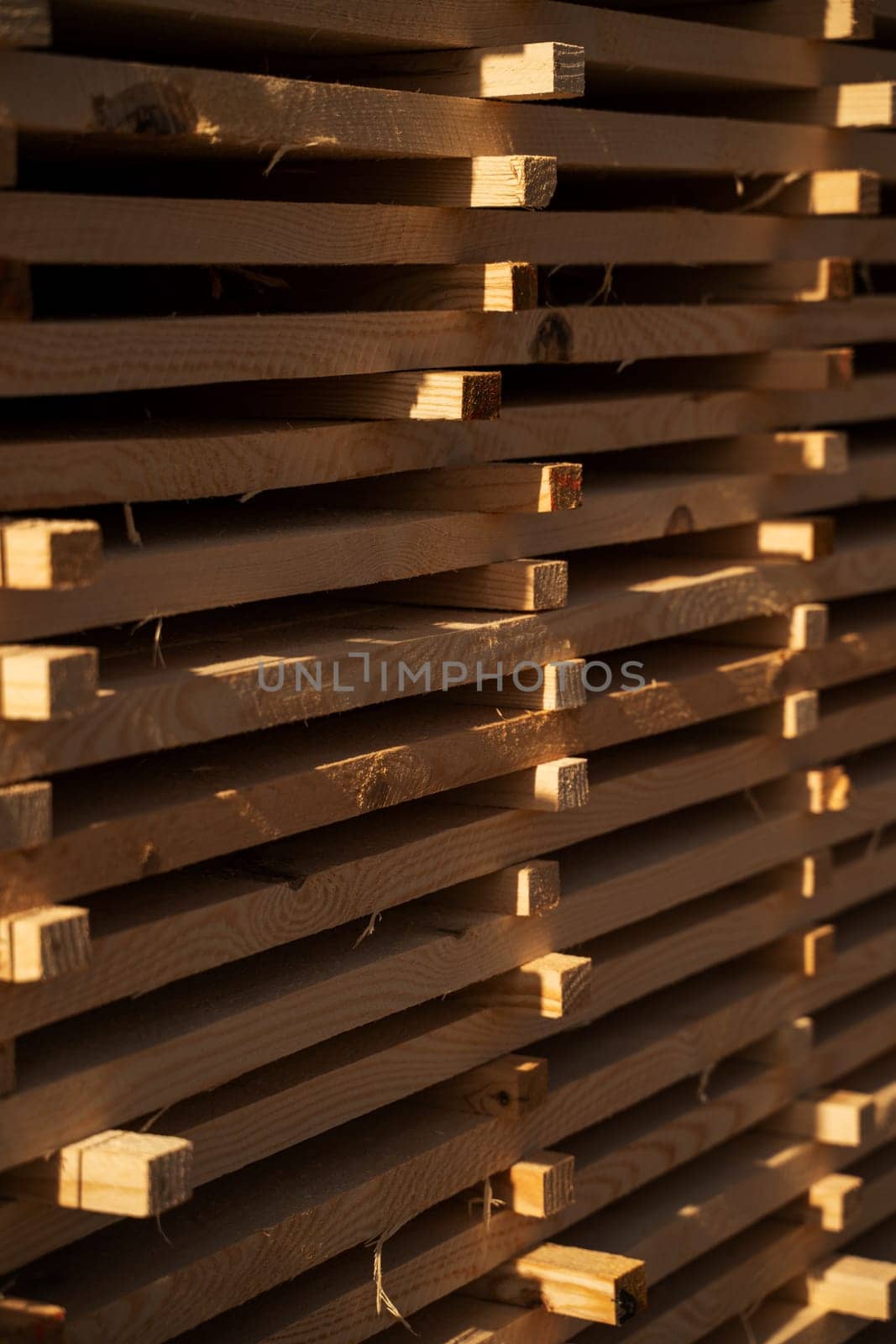 A close-up of neatly stacked wooden boards drying in sunlight at a sawmill. The pattern of the planks creates an industrial texture with rich golden tones.