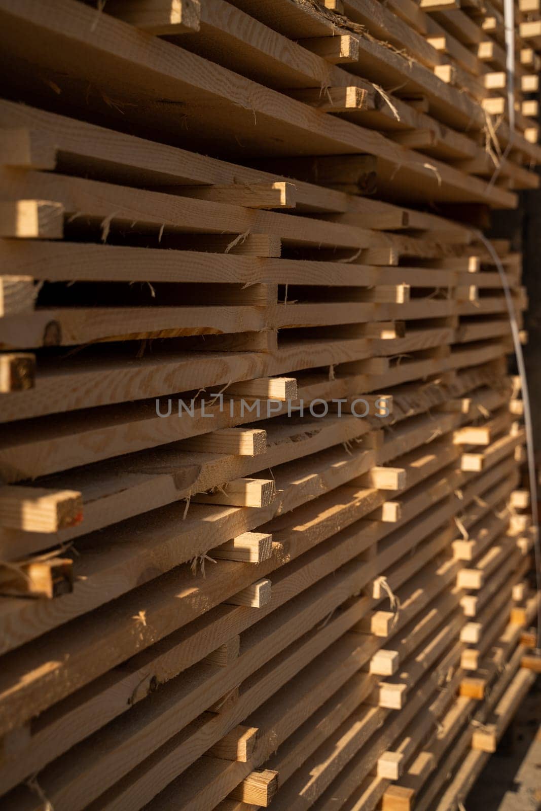 A close-up of neatly stacked wooden boards drying in sunlight at a sawmill. The pattern of the planks creates an industrial texture with rich golden tones.