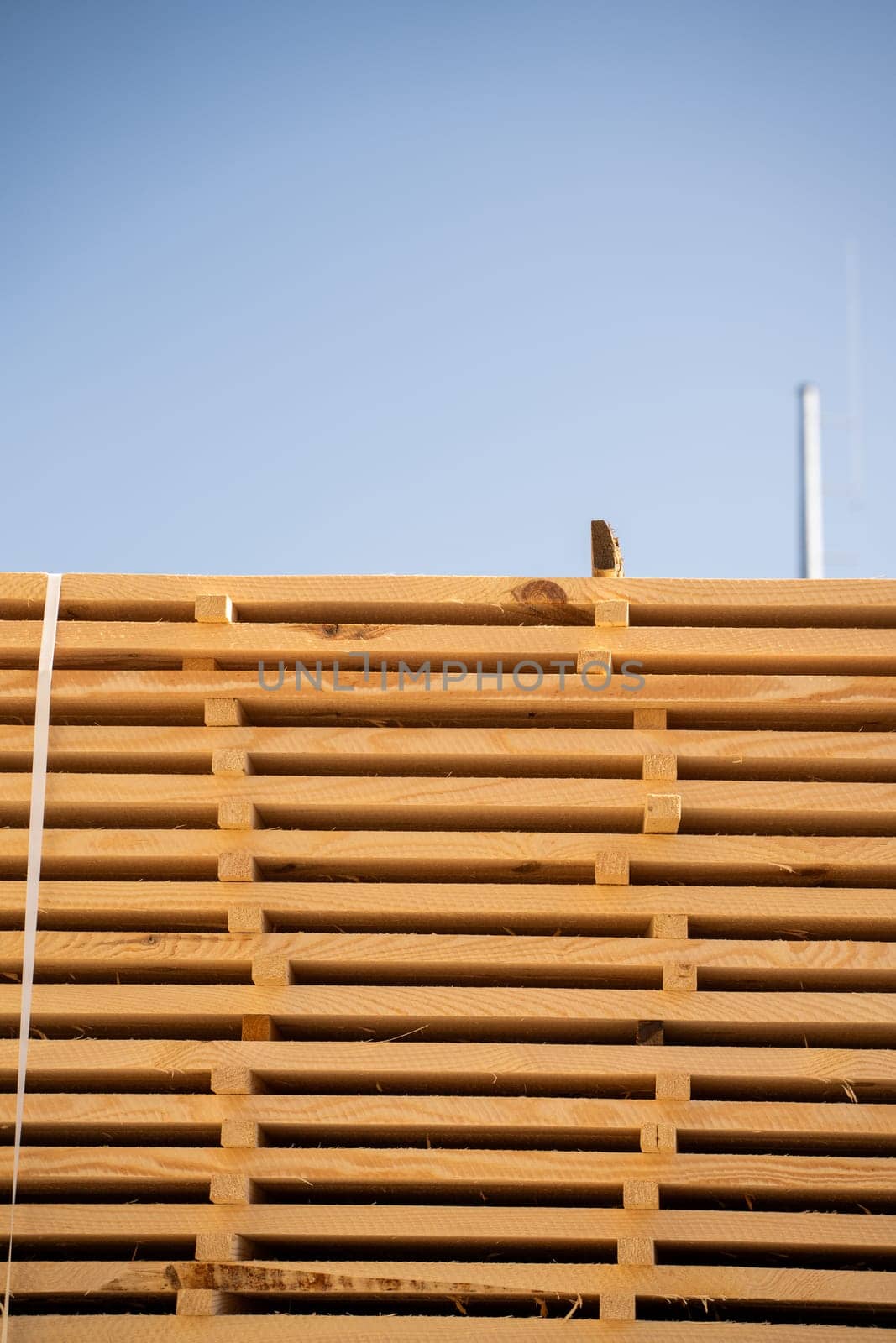 Tall stacks of sawn timber boards drying under open sky next to a building. The light creates strong contrast on the rough, freshly cut wood surfaces.