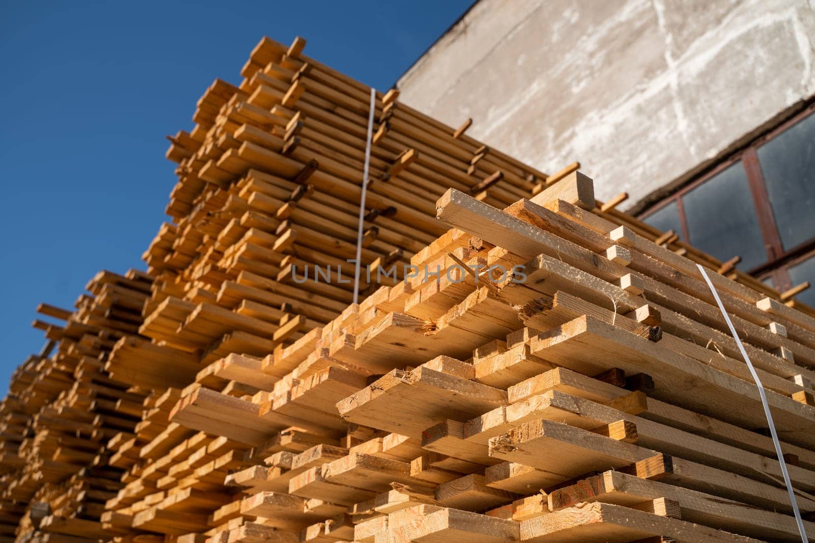 Neatly arranged timber planks are stacked beside an old brick wall, drying in natural light. A vertical plastic strap holds the structure tightly in place.