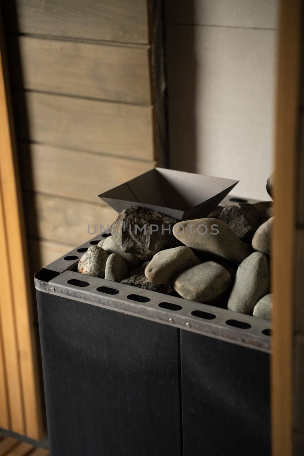 Close view of a metal sauna stove filled with smooth stones, positioned in a wood-paneled relaxation room. Natural light highlights the warm texture and form.