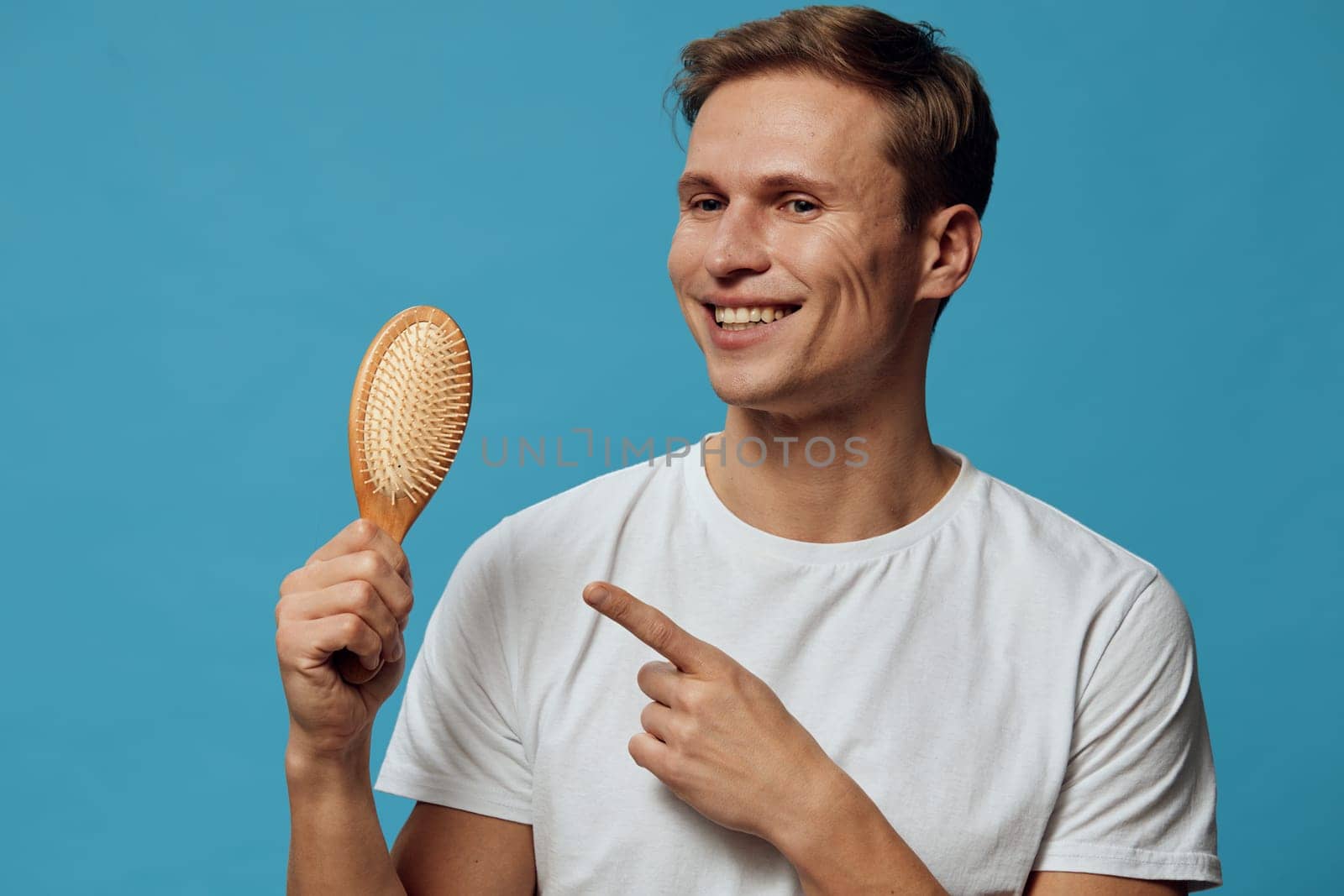 Happy young man with short brown hair and cheerful expression poses against vibrant blue background, holding wooden hairbrush and pointing at it. Concept: personal grooming and self-care, positive attitude, studio portrait.