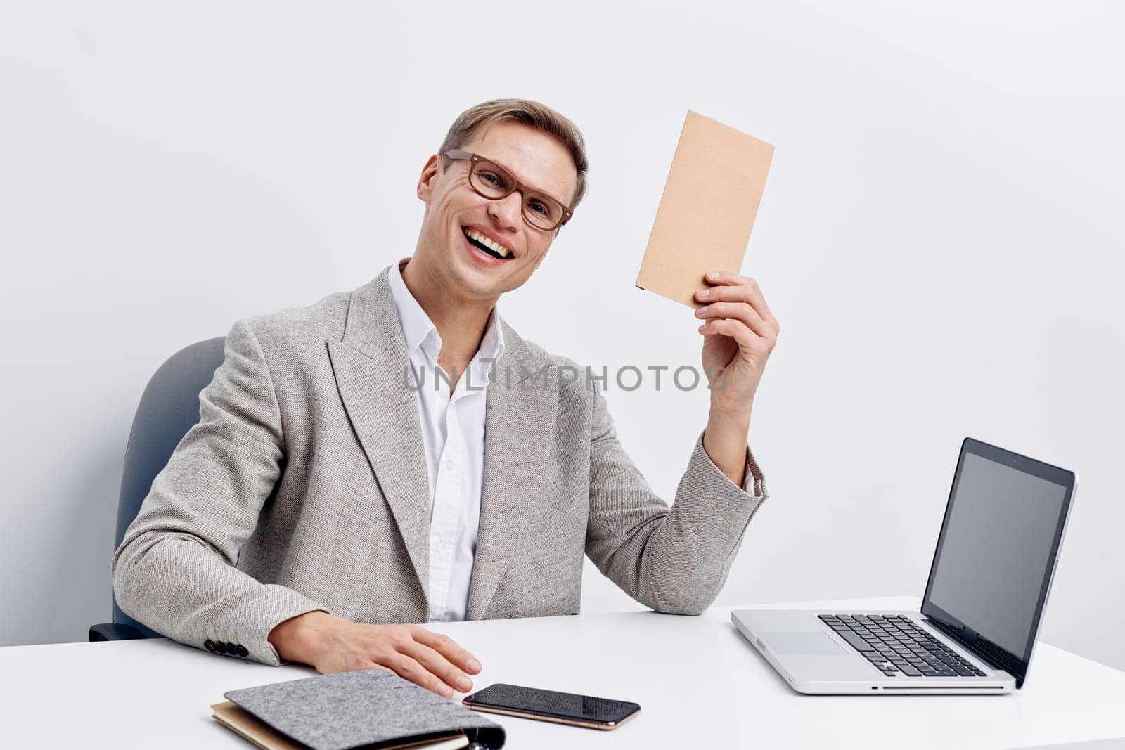smiling man remote work holding envelope sitting at white desk with laptop and smartphone in casual gray jacket. cheerful young caucasian male professional using digital devices for communication and freelance tasks at home office. Concept: digital nomad and communication