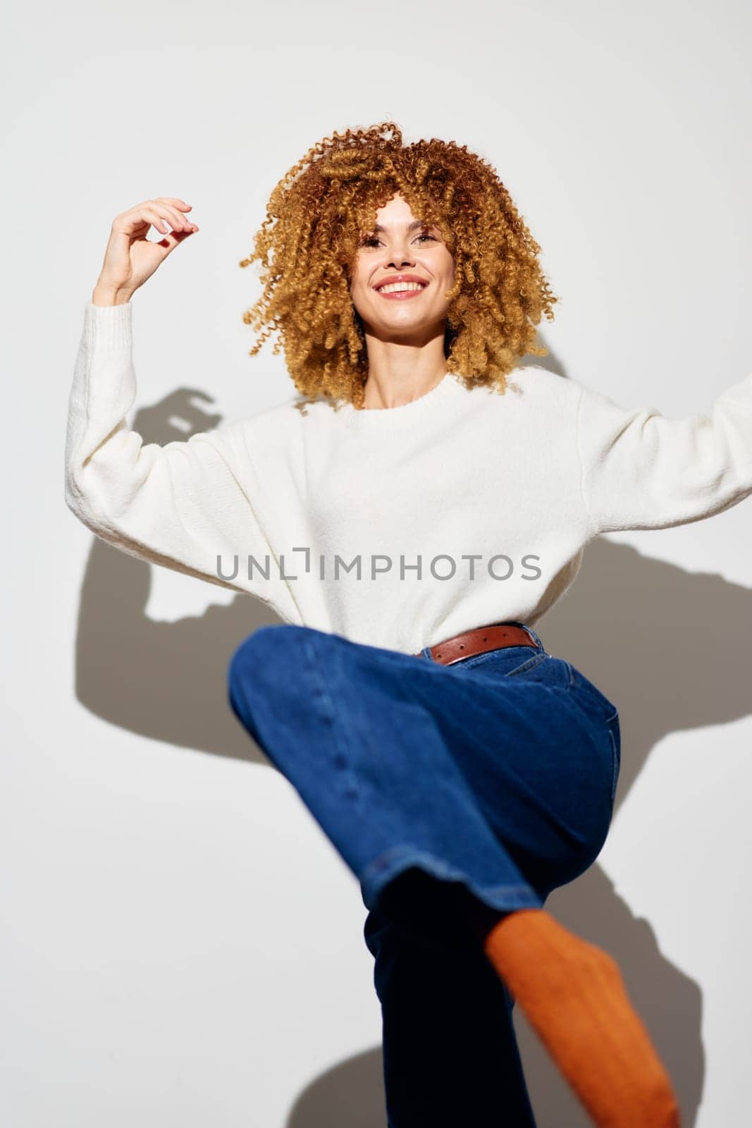 Cheerful woman with voluminous curly hair wearing a cozy white sweater and denim jeans, striking a dynamic pose on a neutral white backdrop, radiating positivity and confidence