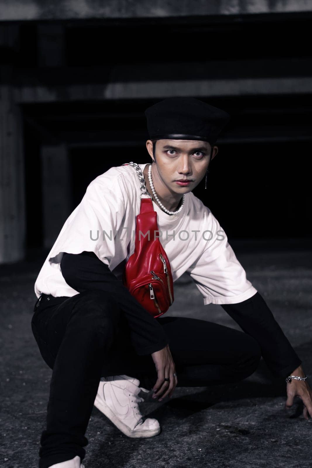 an Asian man sitting around an old building with white clothes and a red bag on his body at night