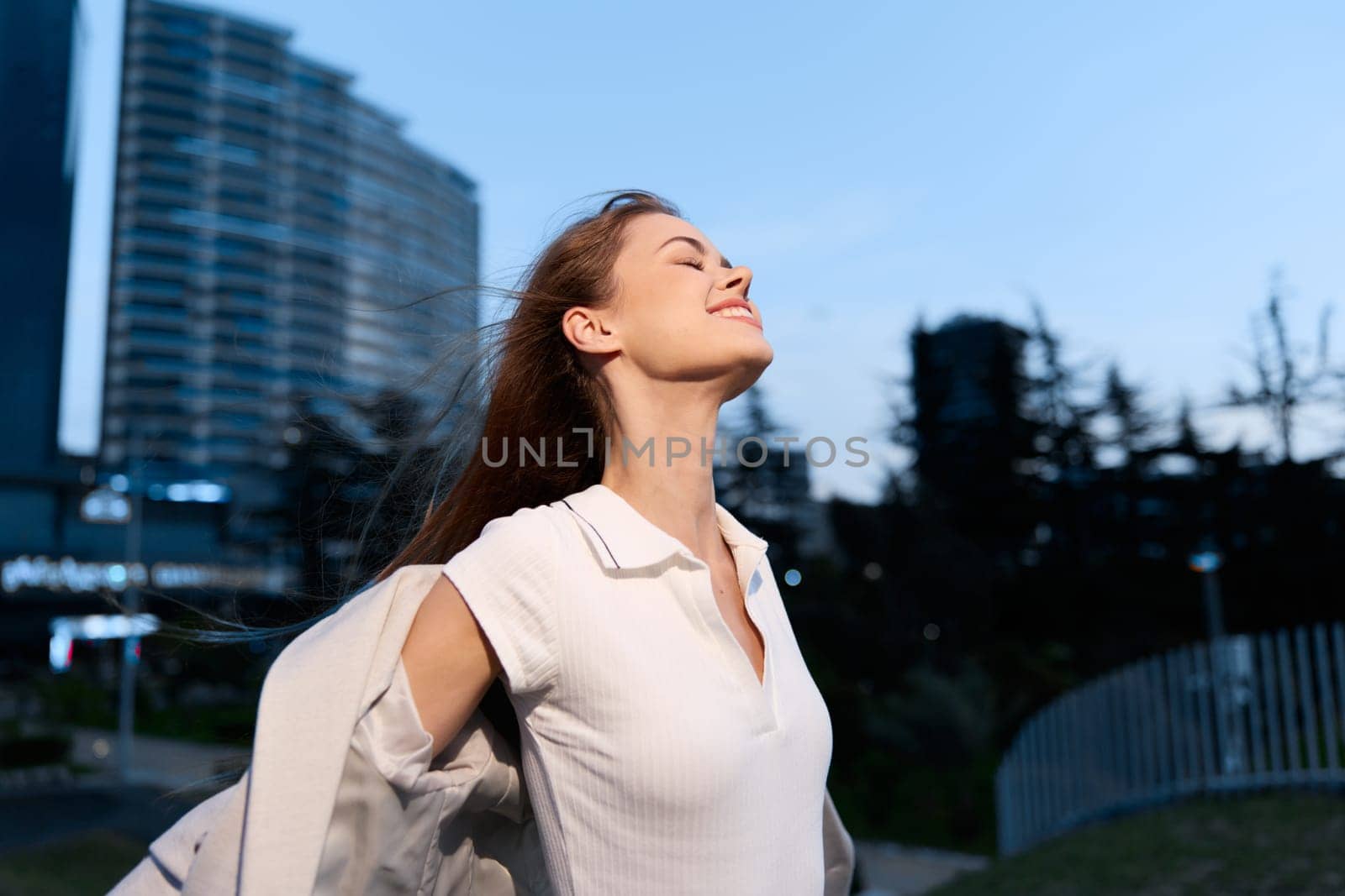 A confident woman exudes positivity and strength in a fashionable outfit, embracing the evening breeze in an urban landscape, symbolizing success and empowerment in business.