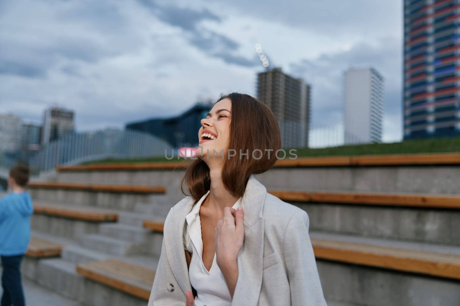 Woman wearing white blouse and light coat laughing joyfully outdoors with urban background, expressing happiness, positivity, and confidence in a business fashion context.