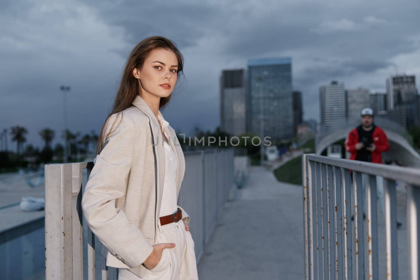 Confident woman in neutral suit poses on urban bridge with city skyline and overcast sky, showcasing fashion style and calm mood in modern outdoor setting