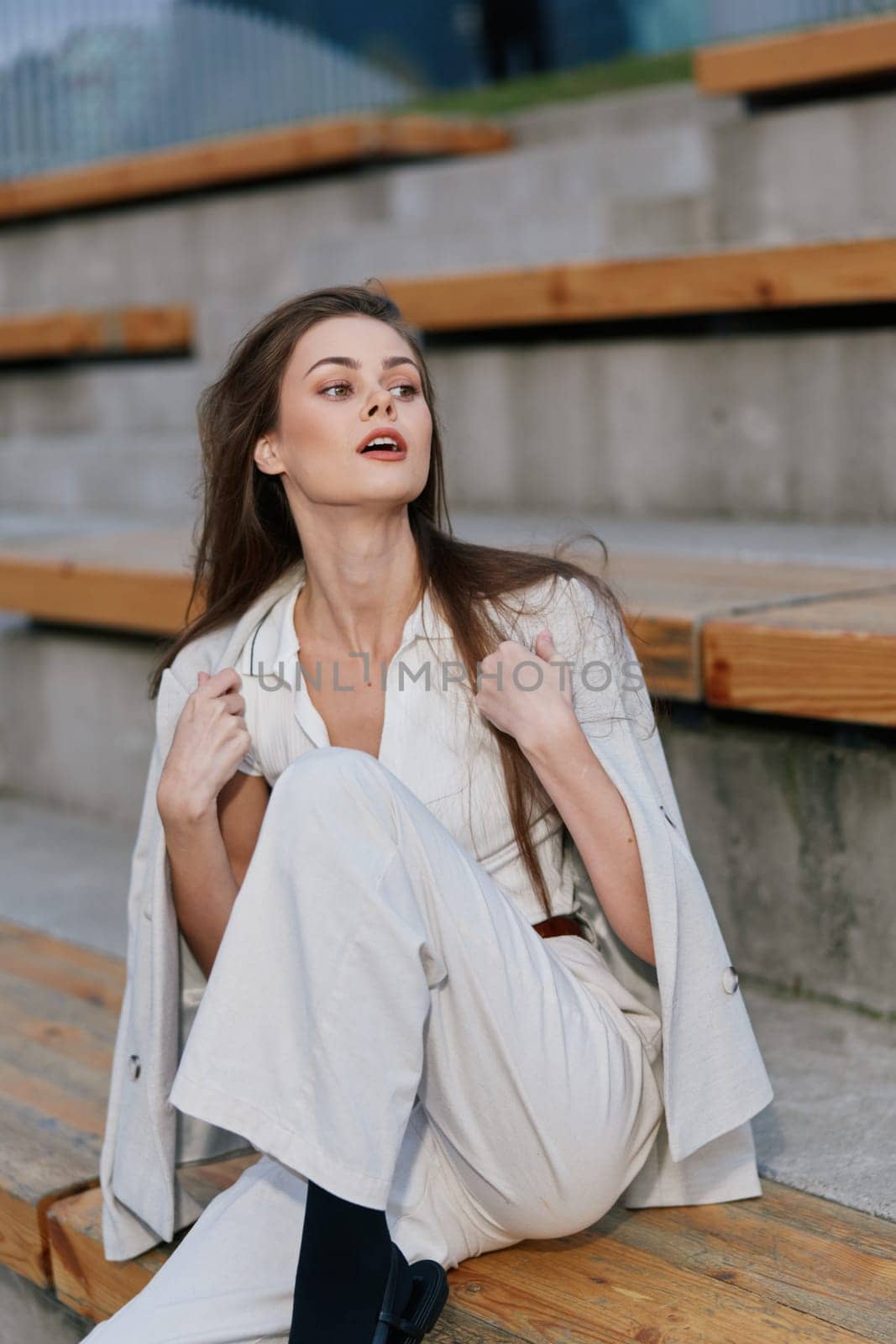 Confident young white woman in elegant white outfit and blazer sits on wooden stairs in neutral gray urban setting, capturing autumn fashion style with calm emotions.