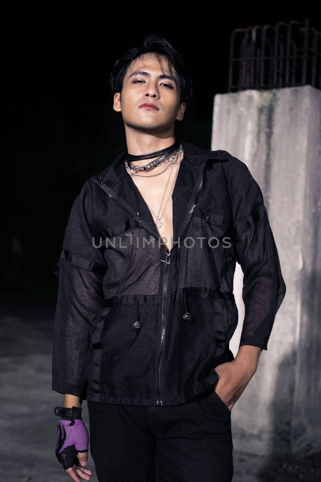 an Asian man dressed all in black and black hair posing as masculine in front of a building pillar