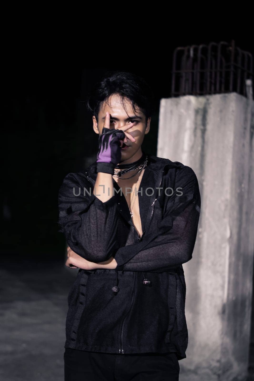 an Asian man dressed all in black and black hair posing as masculine in front of a building pillar