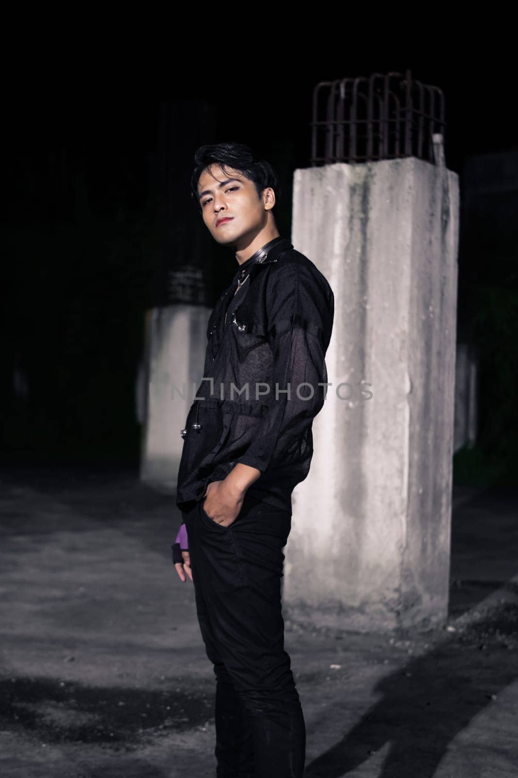 an Asian man dressed all in black and black hair posing as masculine in front of a building pillar