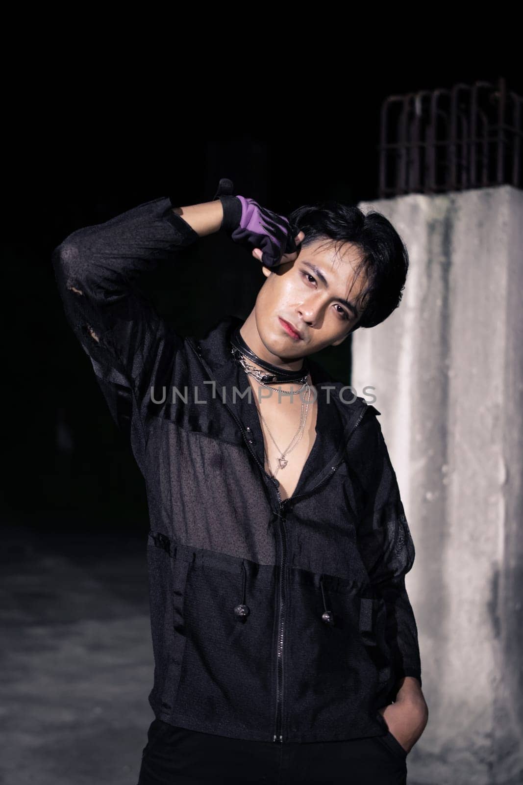 an Asian man dressed all in black and black hair posing as masculine in front of a building pillar