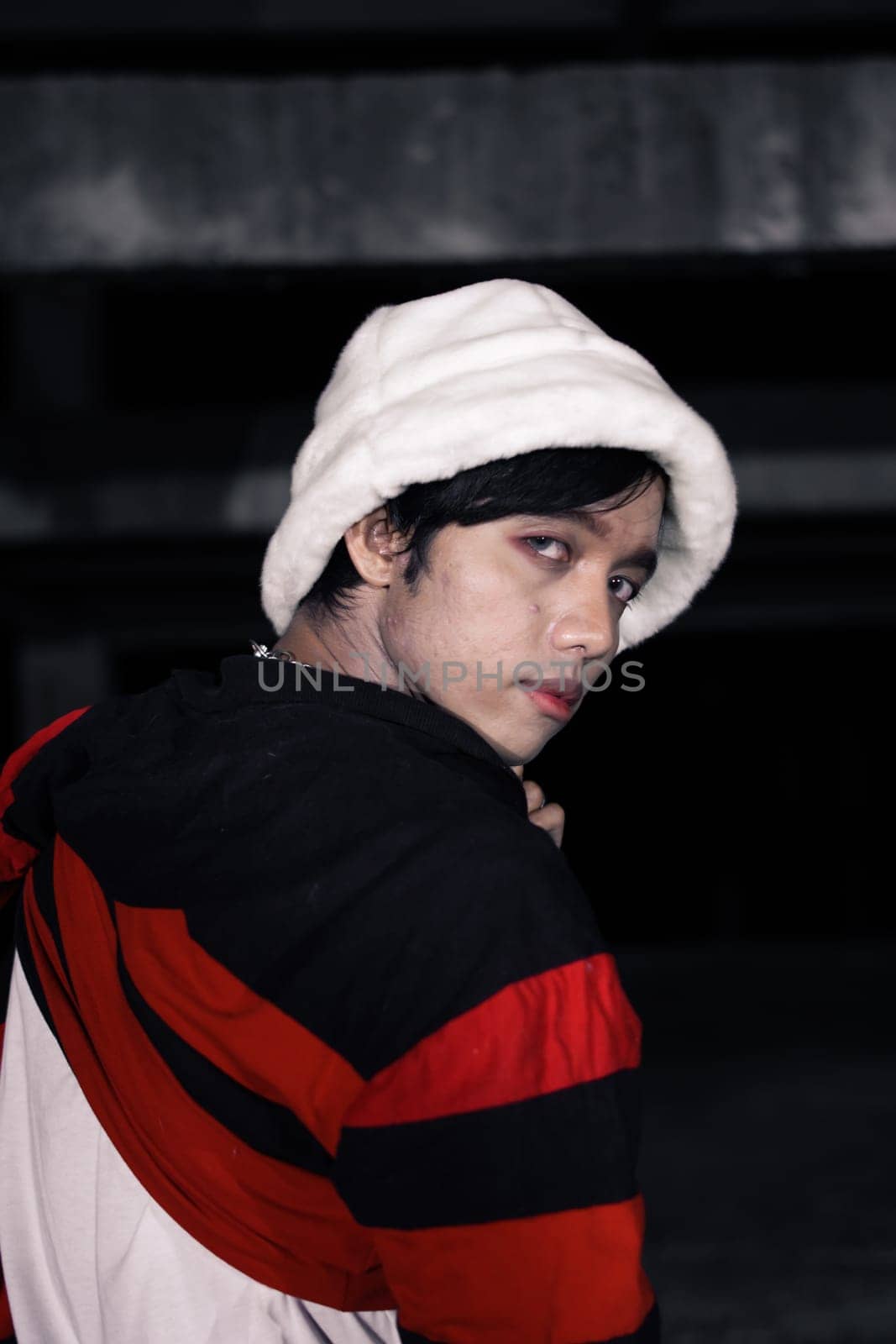 an Asian man in a white velvet hat wears a red sweater and black pants at night