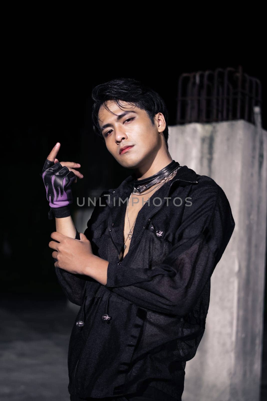 an Asian man dressed all in black and black hair posing as masculine in front of a building pillar