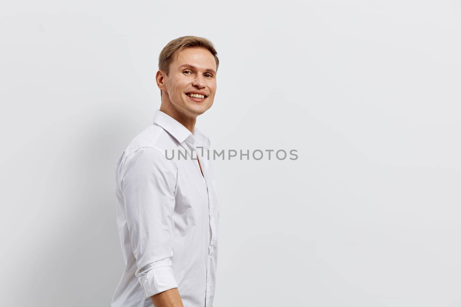 Happy young man with blond hair and blue eyes wearing a white casual shirt, smiling warmly and looking directly at the camera in a bright studio setting with a plain white background. Concept: professional confidence and positivity
