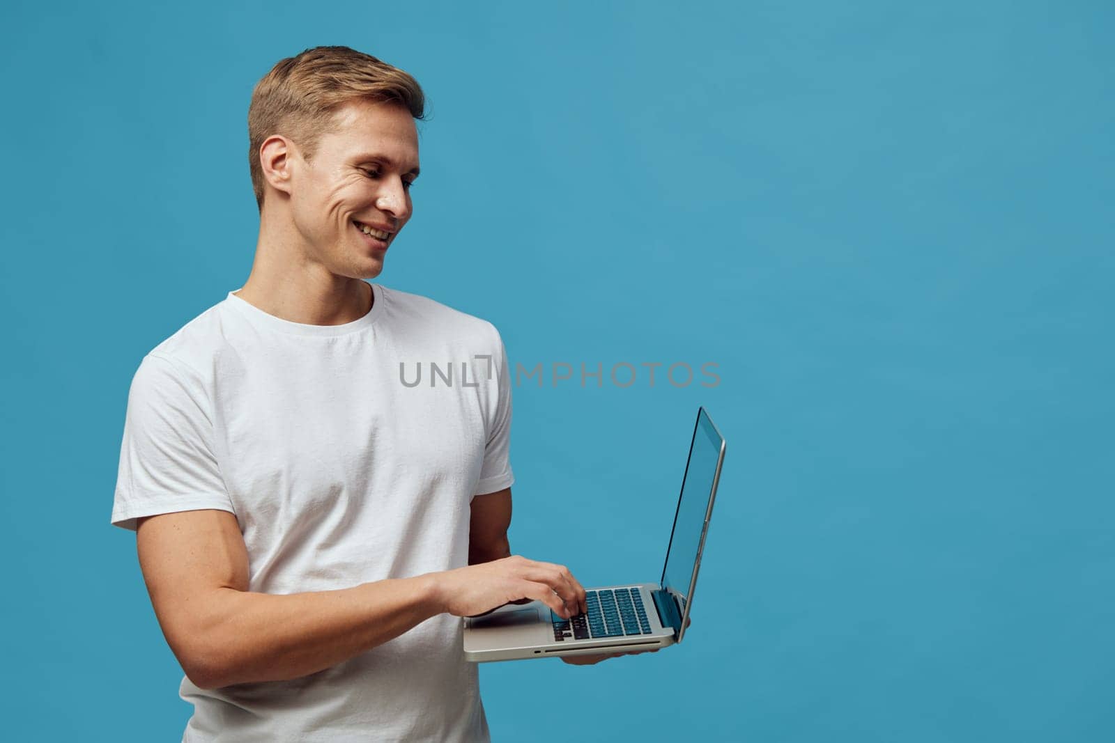 Happy young man wearing a casual white T-shirt, smiling while holding a laptop against a solid blue background. Perfect for technology, remote work, digital communication, future of work, youth lifestyle, casual office setting. Concept: modern tech lifestyle.