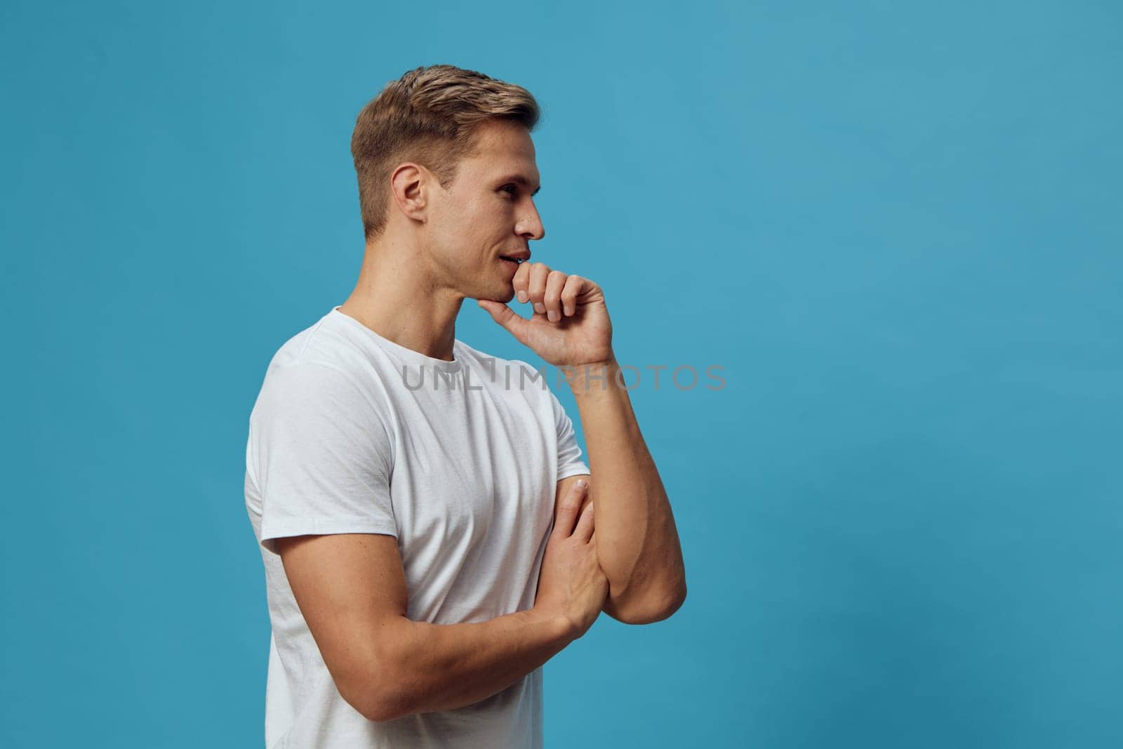 Confident young man in a white casual t-shirt stands with crossed arms and a hand on his chin, contemplating or thinking, with a bright blue background. Concept: relaxed confidence and introspection.