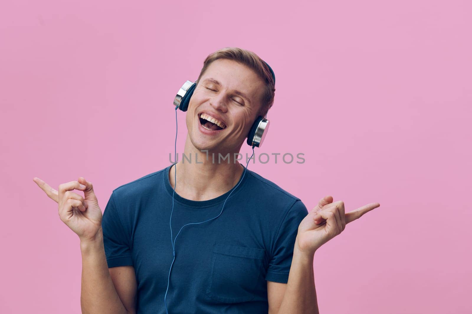 Happy man listening to music with headphones, smiling broadly against pink backdrop, casual attire, carefree attitude, youthful energy, vibrant vibe, lifestyle concept, studio shot.
