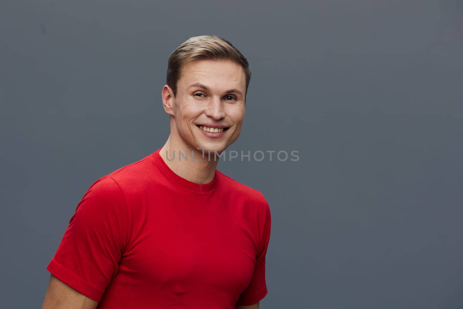 Smiling young caucasian man wear red t-shirt looking directly at camera with friendly expression against gray background in studio portrait style. Concept of virtual communication with digital devices and mental health.