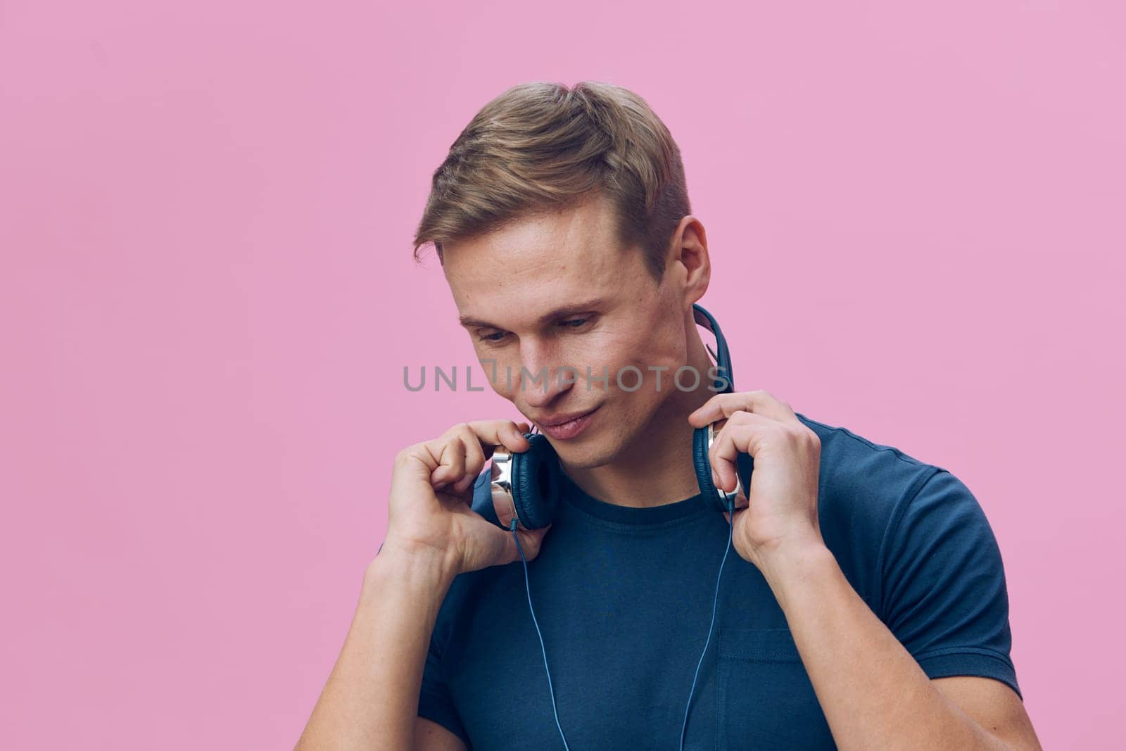Portrait of a cheerful young man with short brown hair, wearing a blue t-shirt and headphones, looking down with a happy expression on a pink studio background, relaxed and modern mood, casual fashion, lifestyle shoot, conceptual image.