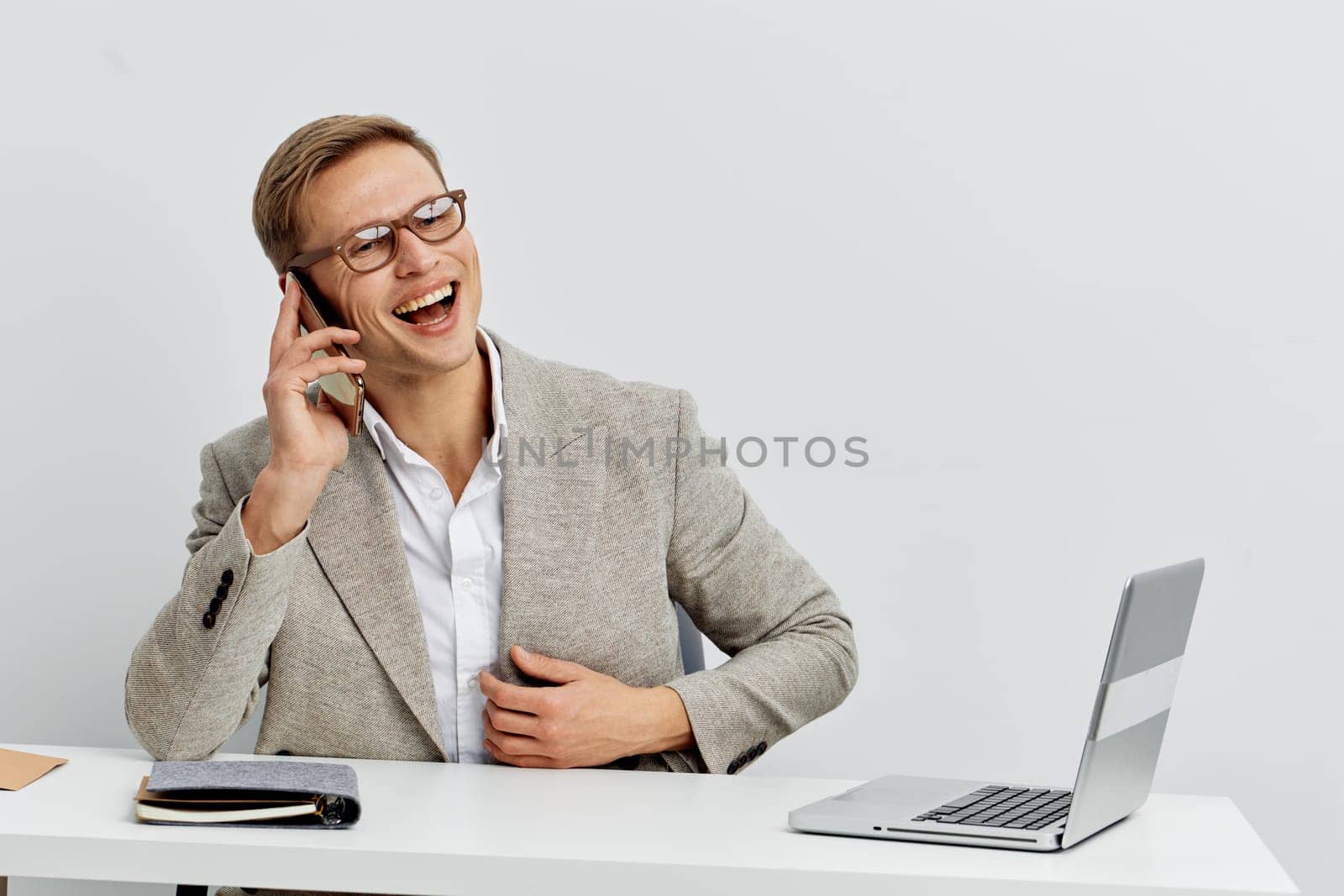 cheerful young man wearing glasses and light gray blazer talking on smartphone and smiling while sitting at white desk with laptop in bright modern space representing digital nomad lifestyle and remote work concept