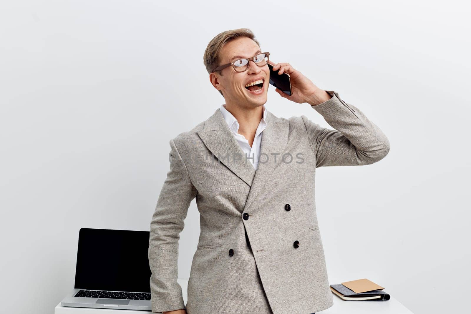 Smiling man dressed in a grey blazer and white shirt using a mobile phone in a professional office setting with a laptop and notebooks on the desk, showing enthusiasm and confidence in a business environment. Concept: successful communication.