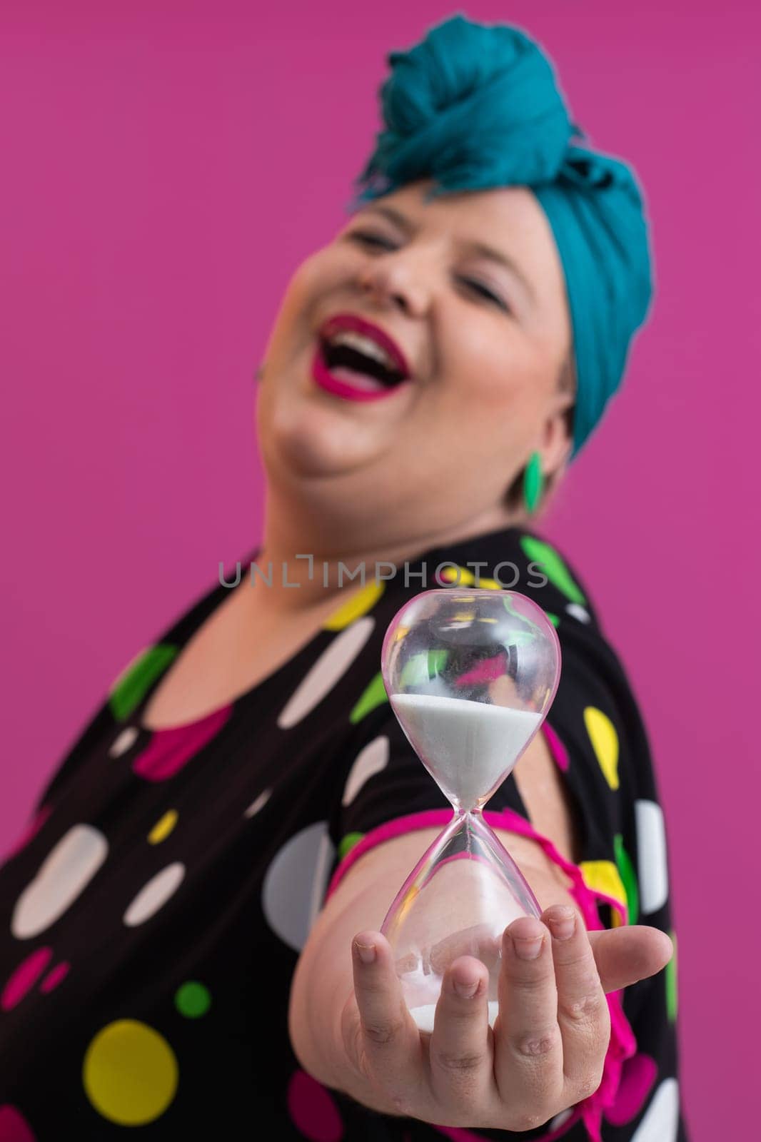 Portrait of a confident woman with a retro headwrap and colorful outfit, holding an hourglass in front of her face as a symbol of time and focus, with a vibrant pink background.