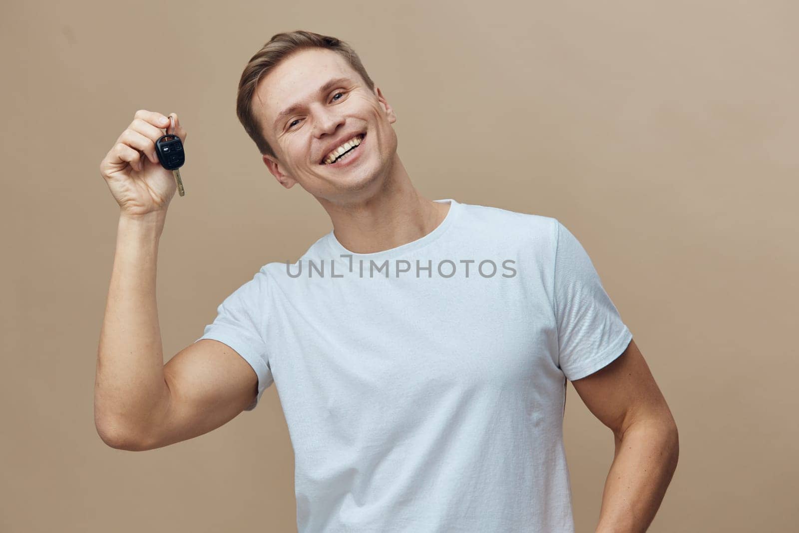 Cheerful man with short brown hair, wearing a light blue t-shirt, displaying a set of clean keys, happy expression, posing against a plain beige studio background, celebrating car ownership, positive emotions.