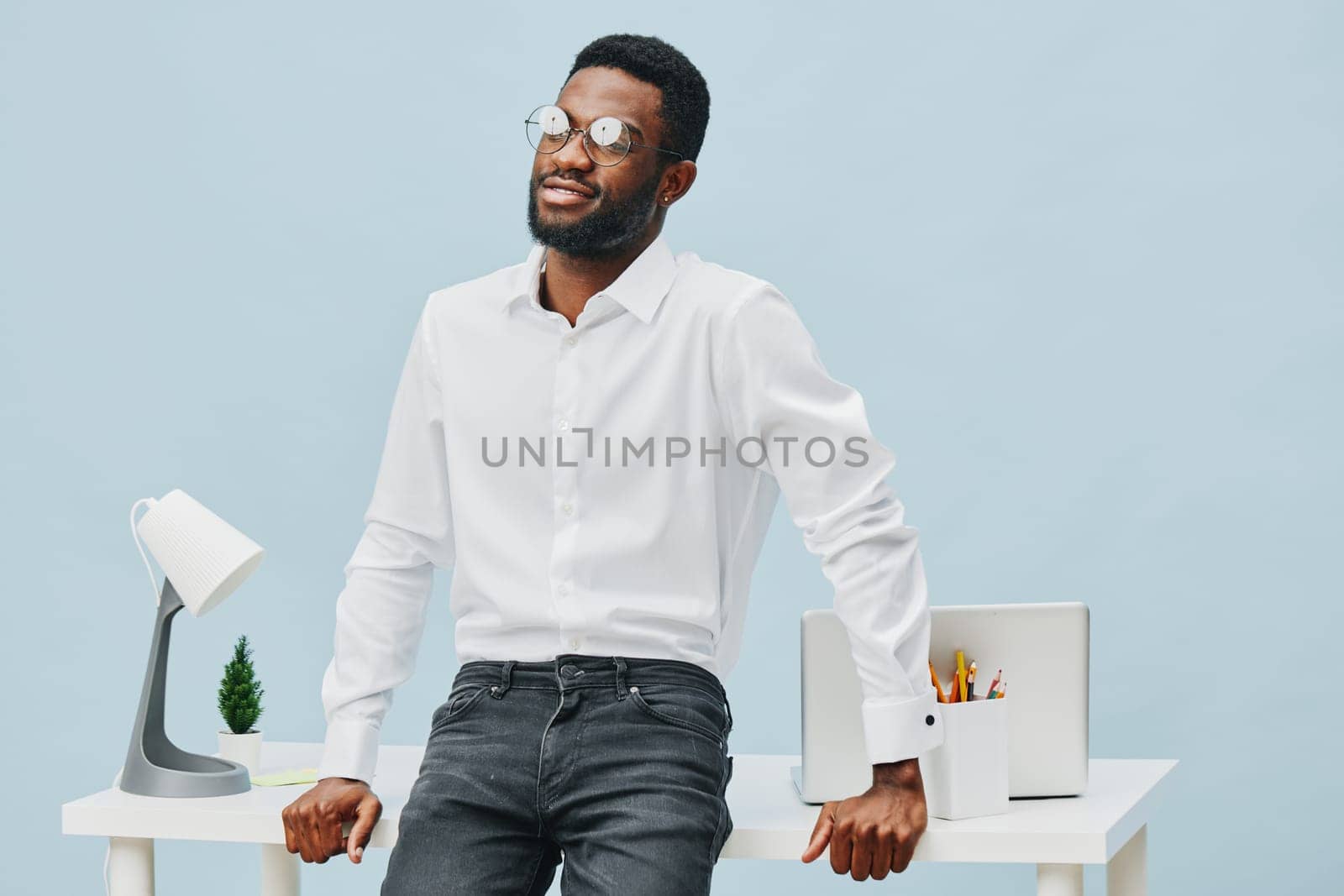 Stylish young Black man dressed in crisp white shirt and dark jeans, posing confidently in a contemporary office setting with a cheerful expression, glasses, and casual professional attire. Bright minimalistic background emphasizes a work and success concept.