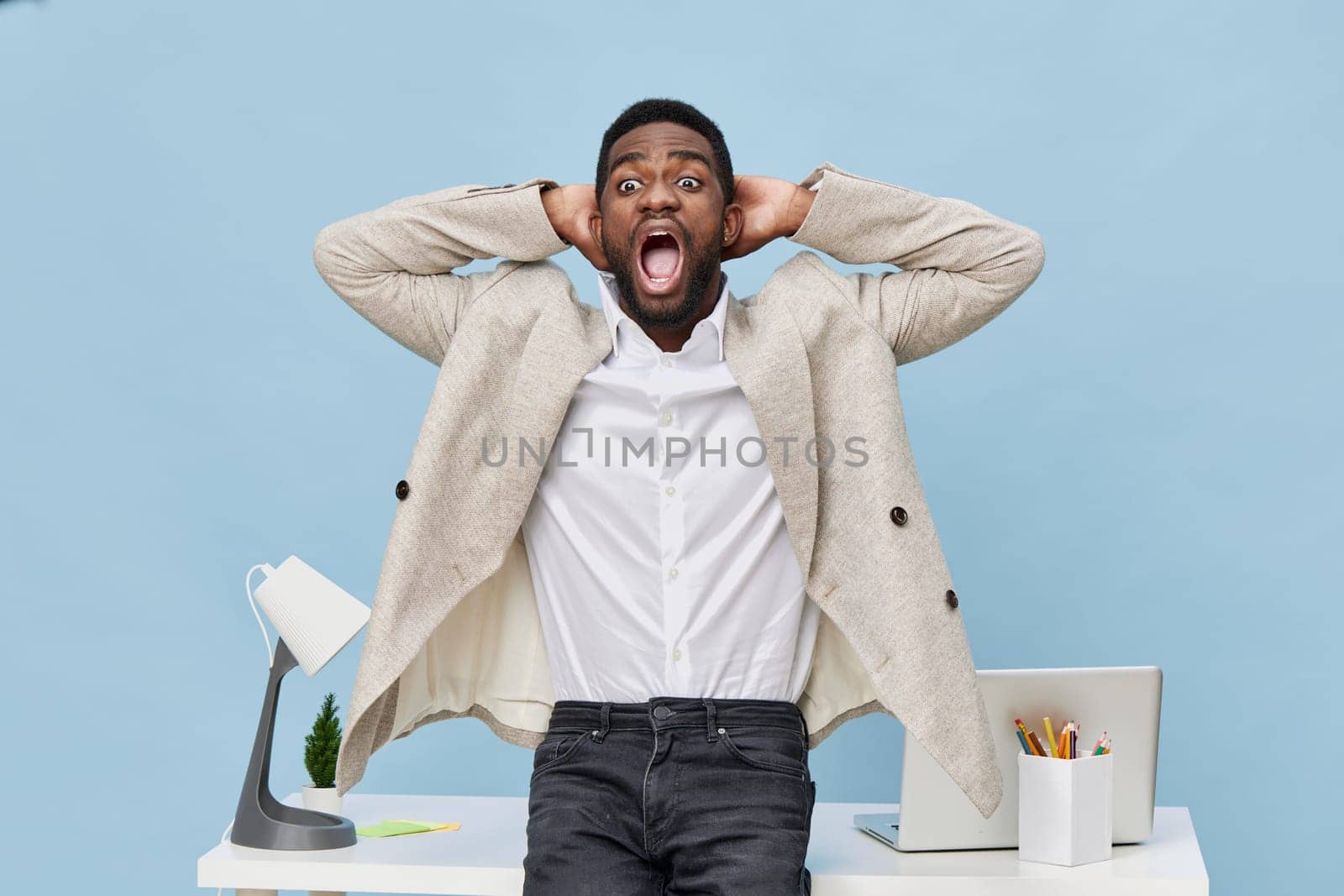 Excited man with youthful appearance, wearing beige blazer and white shirt, expresses shock or frustration at workplace, with hands behind head, in front of cluttered desk, computer, stationery, light blue studio backdrop. Concept: workplace stress and emotions
