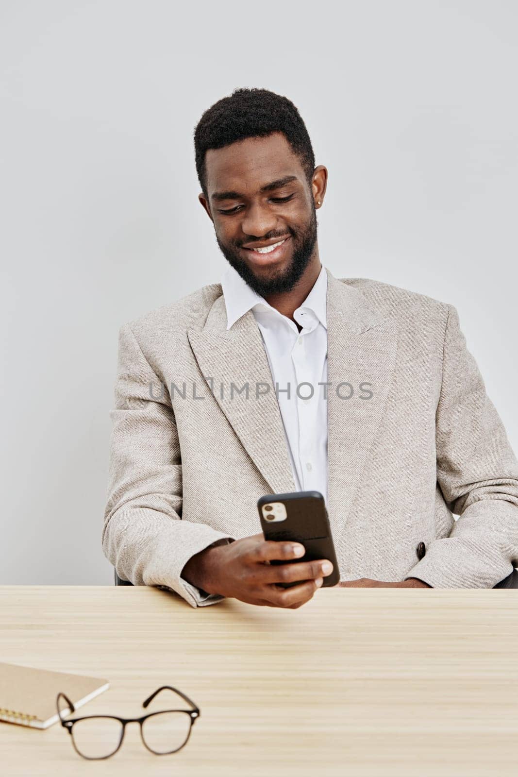 Cheerful man engaged with mobile device, dressed in casual beige blazer and white shirt, demonstrating positive mood, modern workplace setting, indoors, focused on technology use, relaxed atmosphere, lifestyle and communication themes.