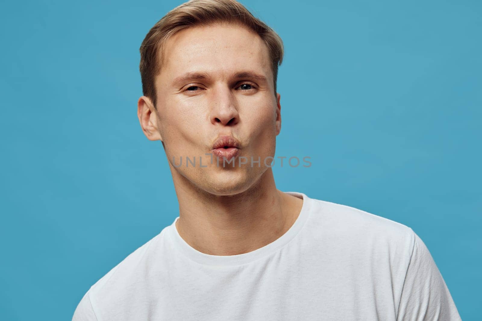 Portrait of a cheerful young man with light brown hair on a vibrant blue background, wearing a white t-shirt, demonstrating a playful and expressive mood, suitable for lifestyle, social media, or youth-themed projects.