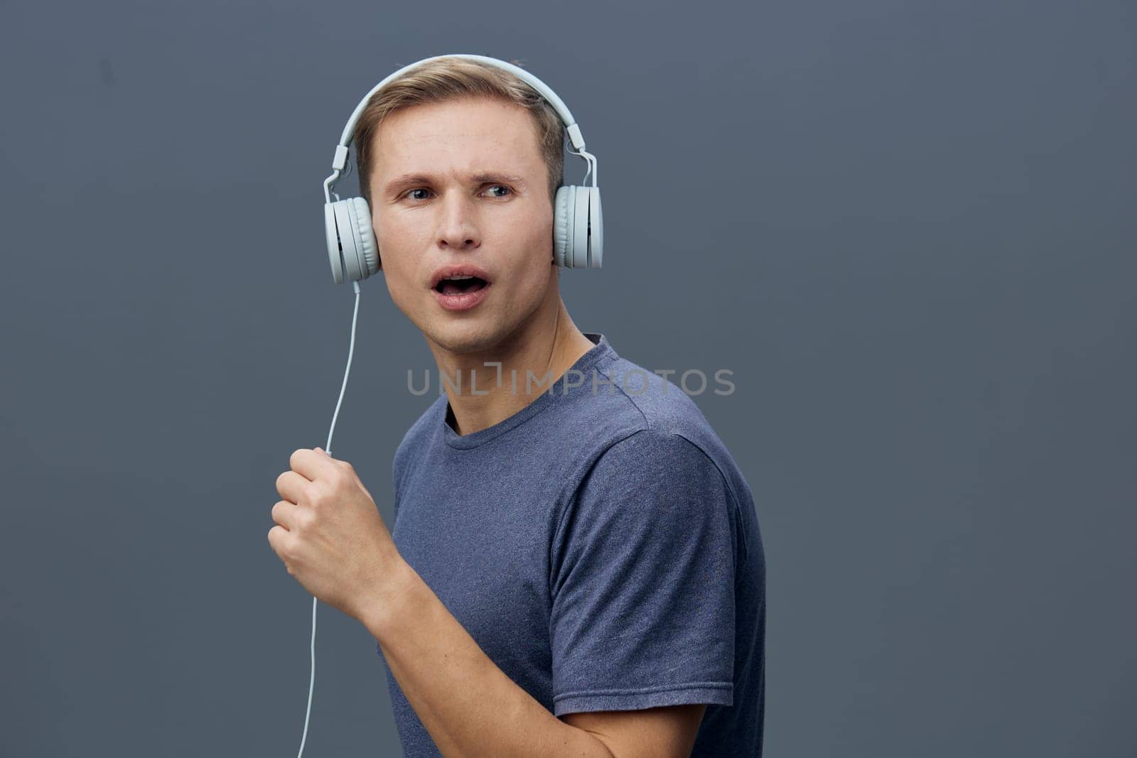 Young man with short hair and fair skin enjoying music through big headphones, expressing surprise or confusion, casual outfit, gray background, studio shot, emphasizing entertainment or sound experience.