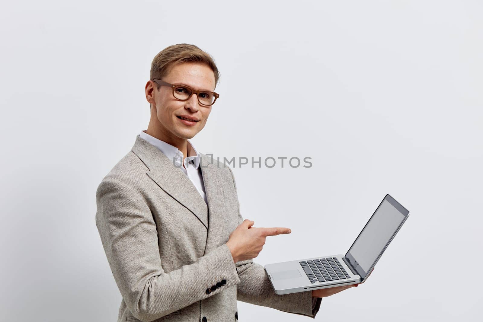 Confident young man wearing formal suit and glasses holding and pointing to open laptop on a plain white background. Business, technology, professional, presentation, communication, success concepts.