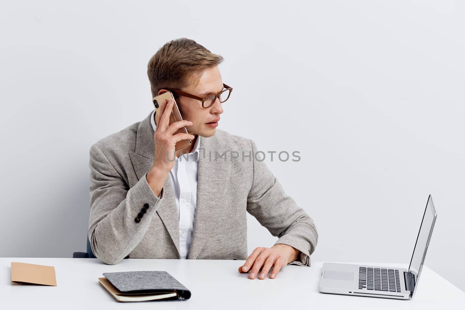 Young professional man with glasses, dressed in a gray blazer and white shirt, sitting at a clean desk using a laptop, talking on a smartphone, and writing notes, isolated on white background. Business, communication, work life