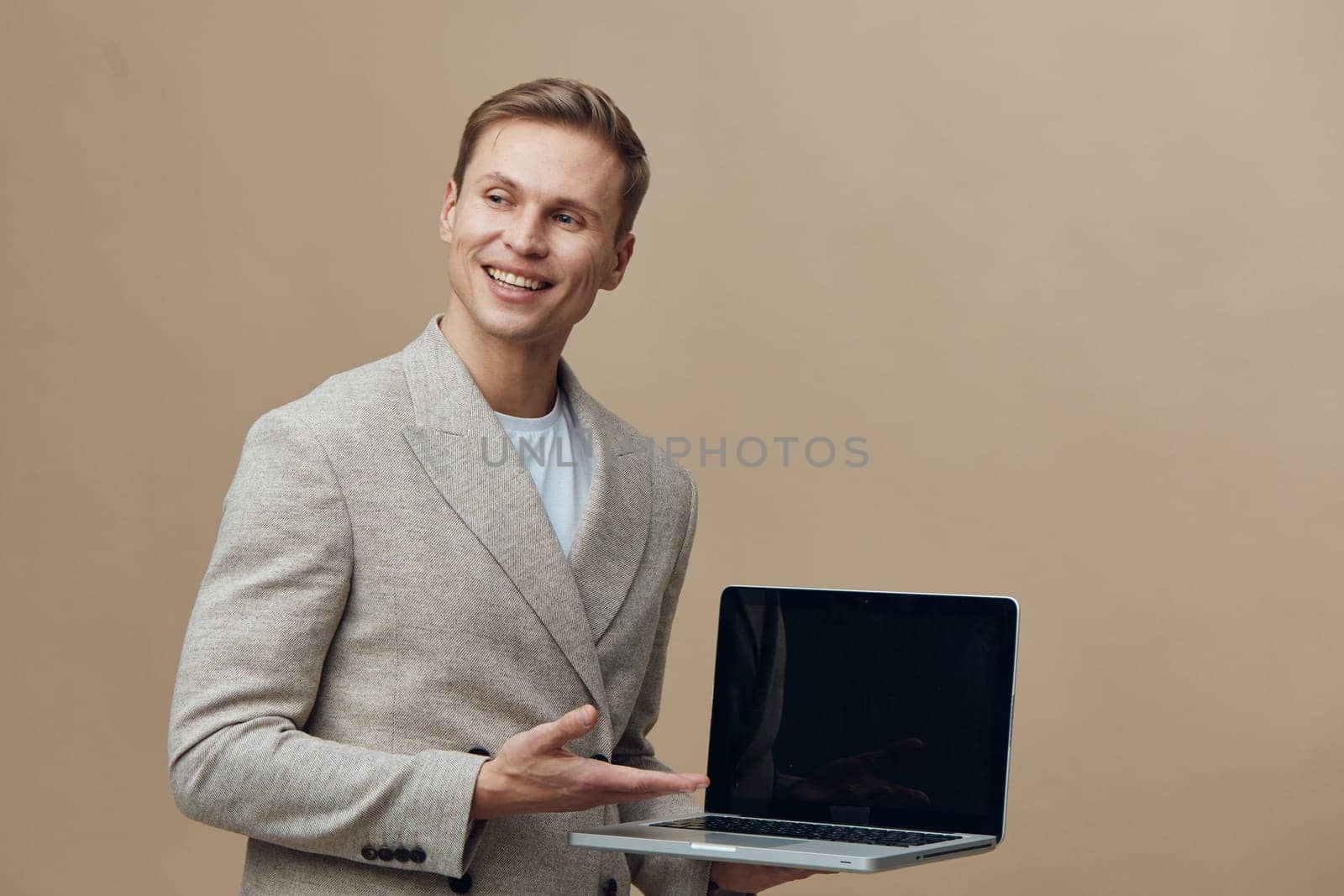 Cheerful young man with bright smile presenting open laptop computer while standing against plain beige backdrop. Experienced in freelance work, technology enthusiast, casual style, modern professional, youthful energy, digital tools, positive attitude. Concept: modern remote work